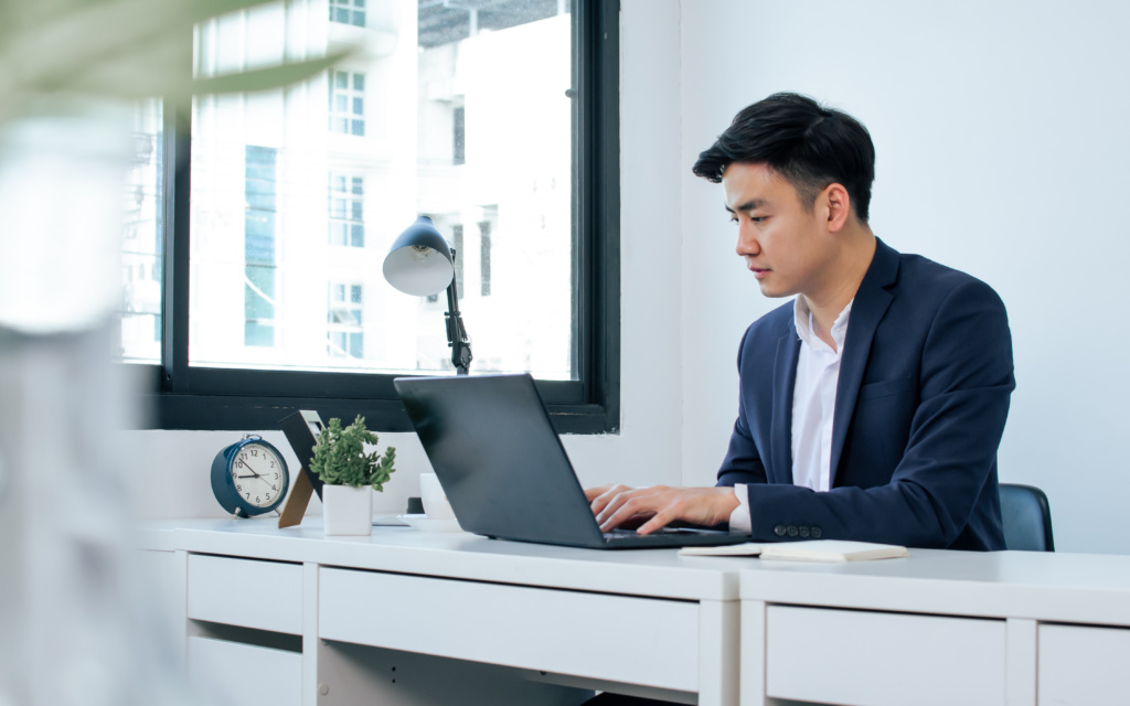 Asian Handsome Business Man Wearing White Shirt And Blue Formal Suit, Communicating, Working, Typing On Laptop, Sitting Near Window In Indoor Office And Smiling With Happiness.