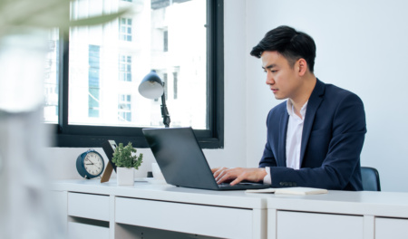 Asian Handsome Business Man Wearing White Shirt And Blue Formal Suit, Communicating, Working, Typing On Laptop, Sitting Near Window In Indoor Office And Smiling With Happiness.